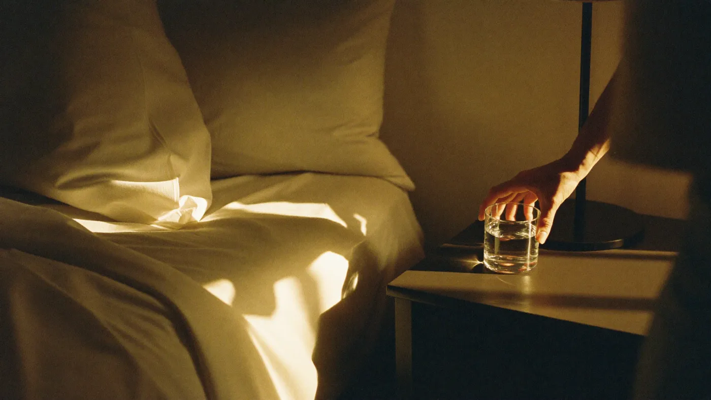 Morning bedroom with sunlight across the sheets and a person's hand reaching for a glass of water on the nightstand