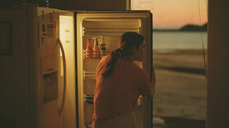 Person standing in front of an open refrigerator at dusk with one hand on the door, tired but calm, warm kitchen light