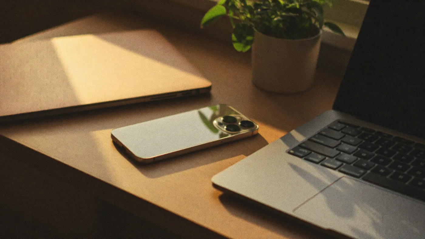Phone face-down on a windowsill beside a small potted plant while a person reads a paper book softly out of focus behind it