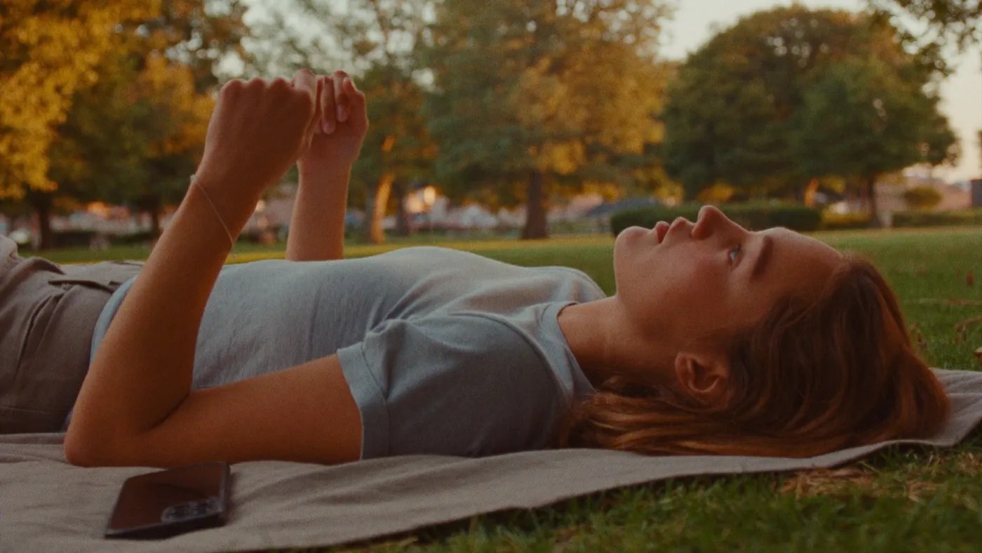 Person setting a phone down on a picnic blanket in a park and looking up at the evening sky, relaxed, golden light
