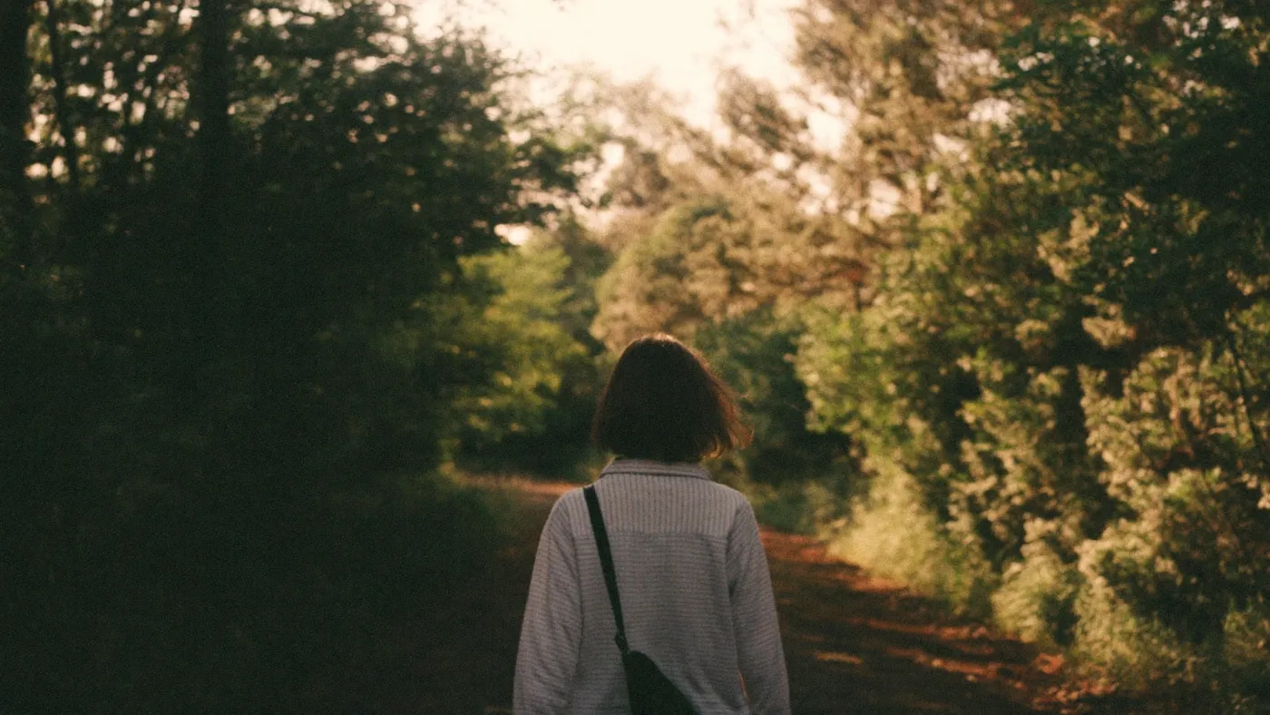 Person walking a quiet forest path with dappled golden light, seen from behind, peaceful and unhurried