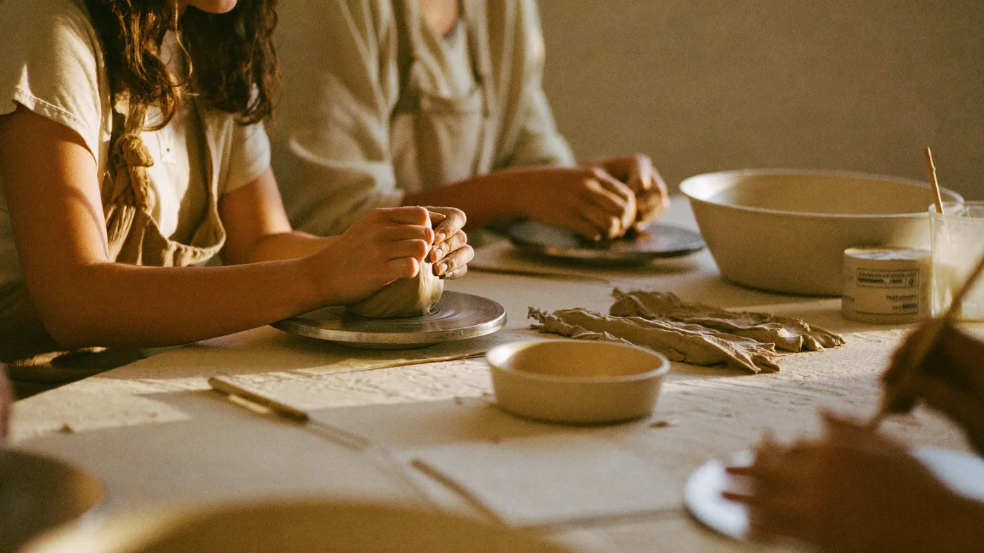 A pottery class table with several pairs of hands shaping clay, warm workshop light, candid