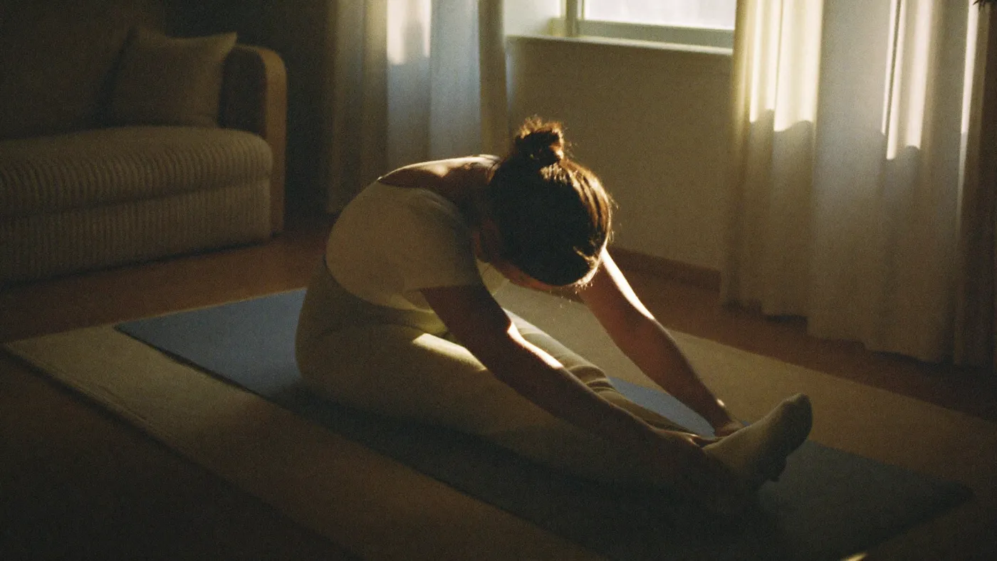 Person in comfortable clothes doing a gentle seated stretch on a yoga mat in a living room, morning light through linen curtains