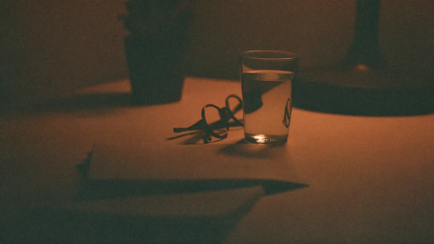 Minimal bedside scene with a glass of water, a small plant, and folded reading glasses in dim warm lamp light