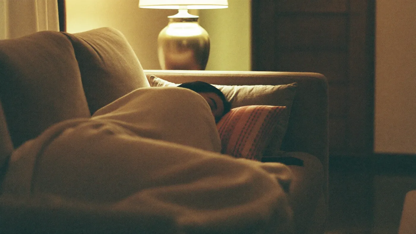 Person resting on a couch under a soft knit blanket, phone face-down on the side table, serene evening lamp light