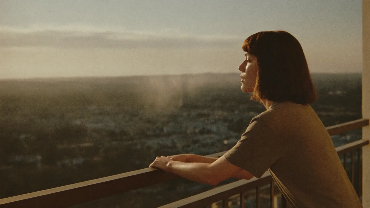Person breathing deeply on a small balcony at sunset, hands on the railing, city softly blurred below