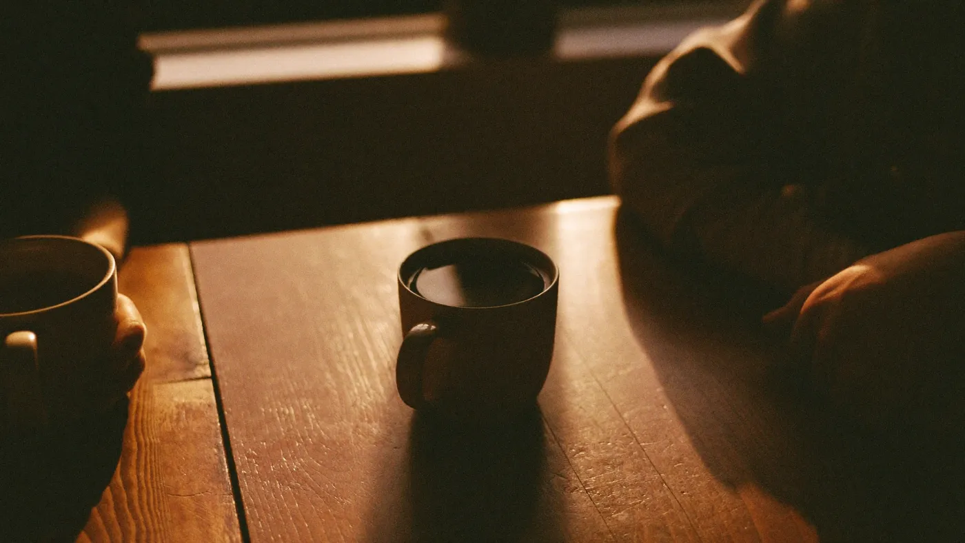 Two mugs of tea on a wooden table between two people in quiet conversation, only hands visible, warm window light