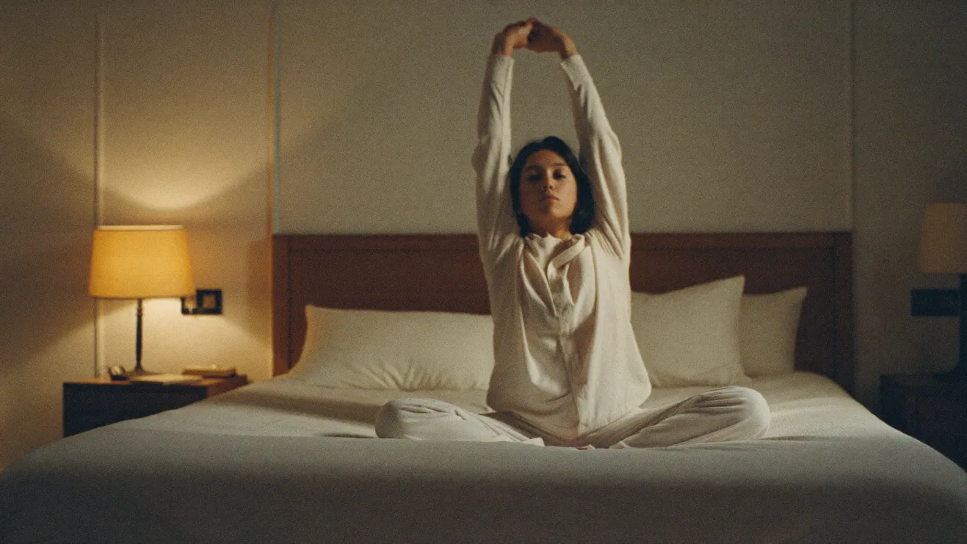 Person sitting on the edge of a bed in soft lamplight, gently stretching, calm expression