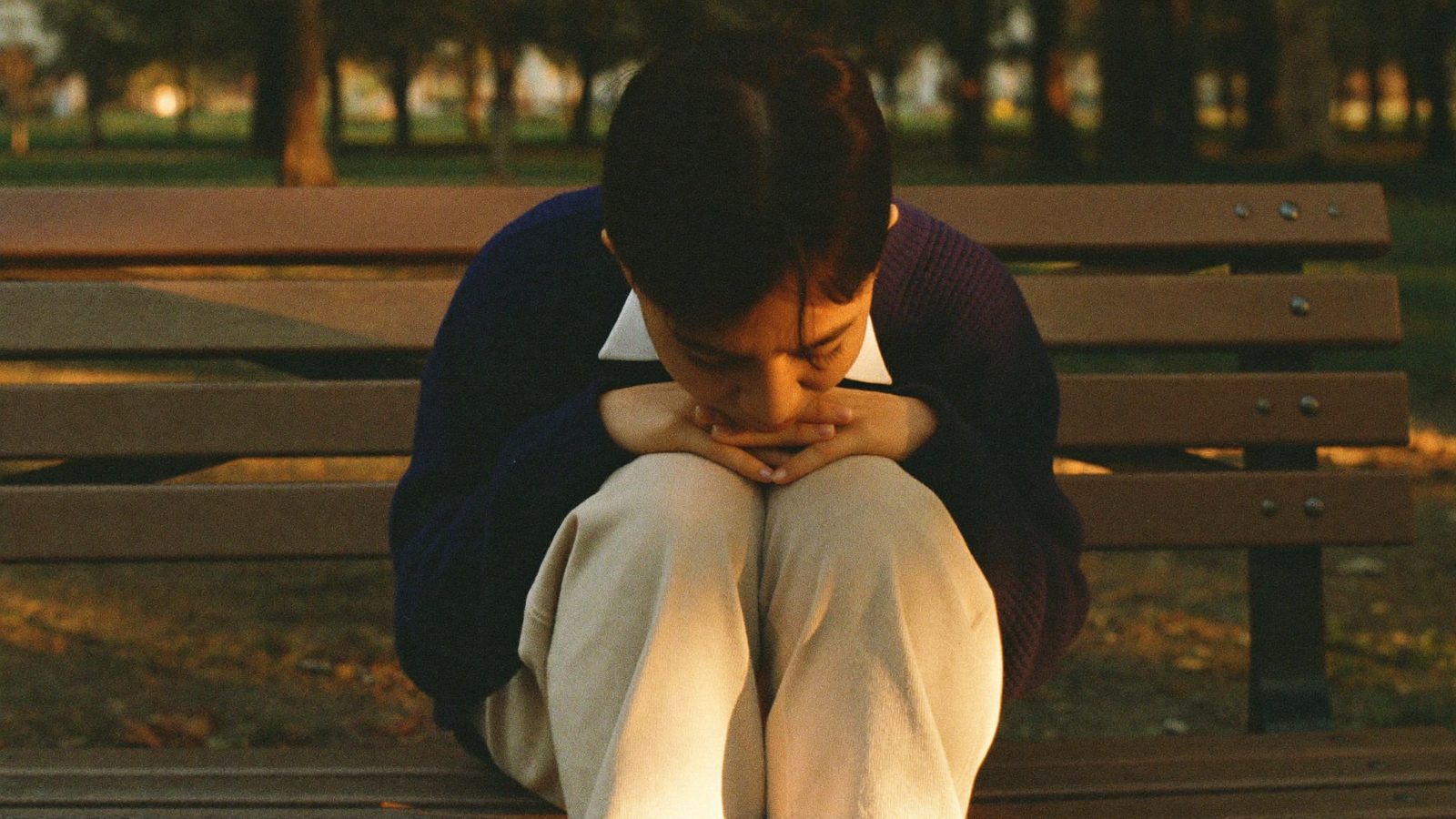 Person sitting on a park bench with both hands flat on their knees, breathing slowly with eyes closed, late golden light through trees