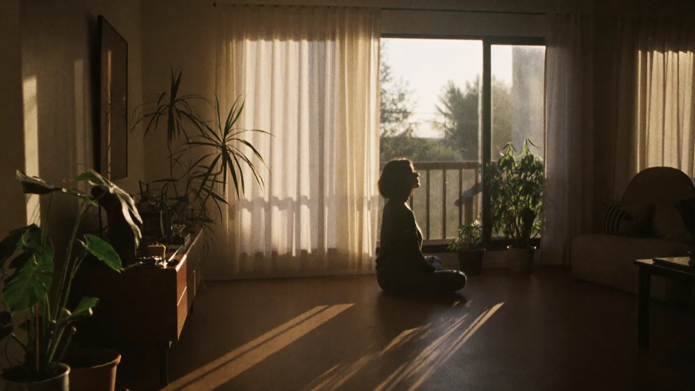 Wide calm living room at golden hour, person seated cross-legged near the window among houseplants, deep serenity