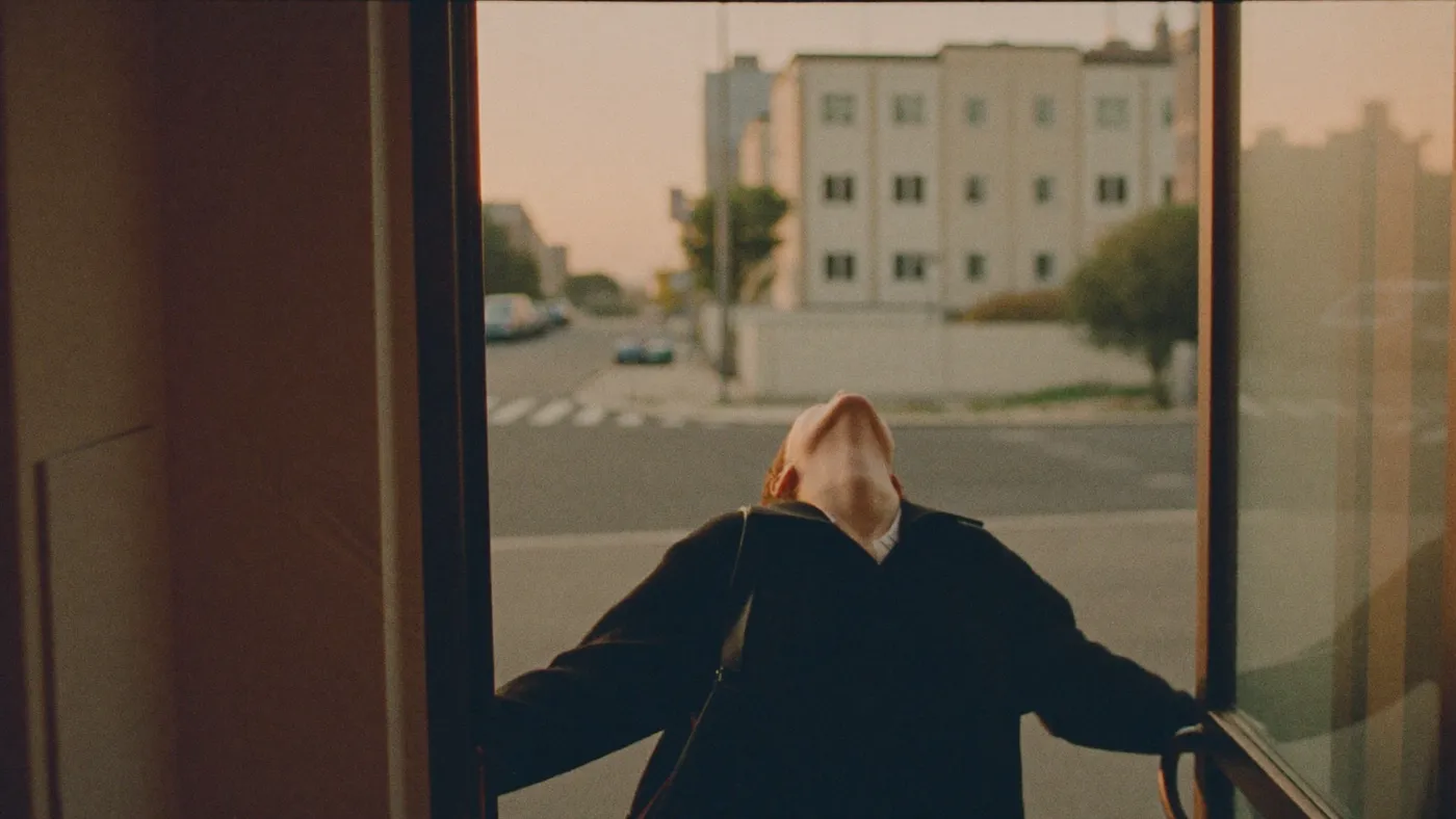 Person stepping out of an office building doorway onto a quiet street at golden hour, shoulders relaxing, relieved exhale