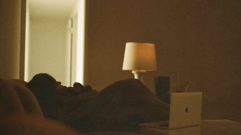 Person on a sofa late at night under a blanket, laptop closed on the coffee table, looking toward a dim hallway, soft lamp light