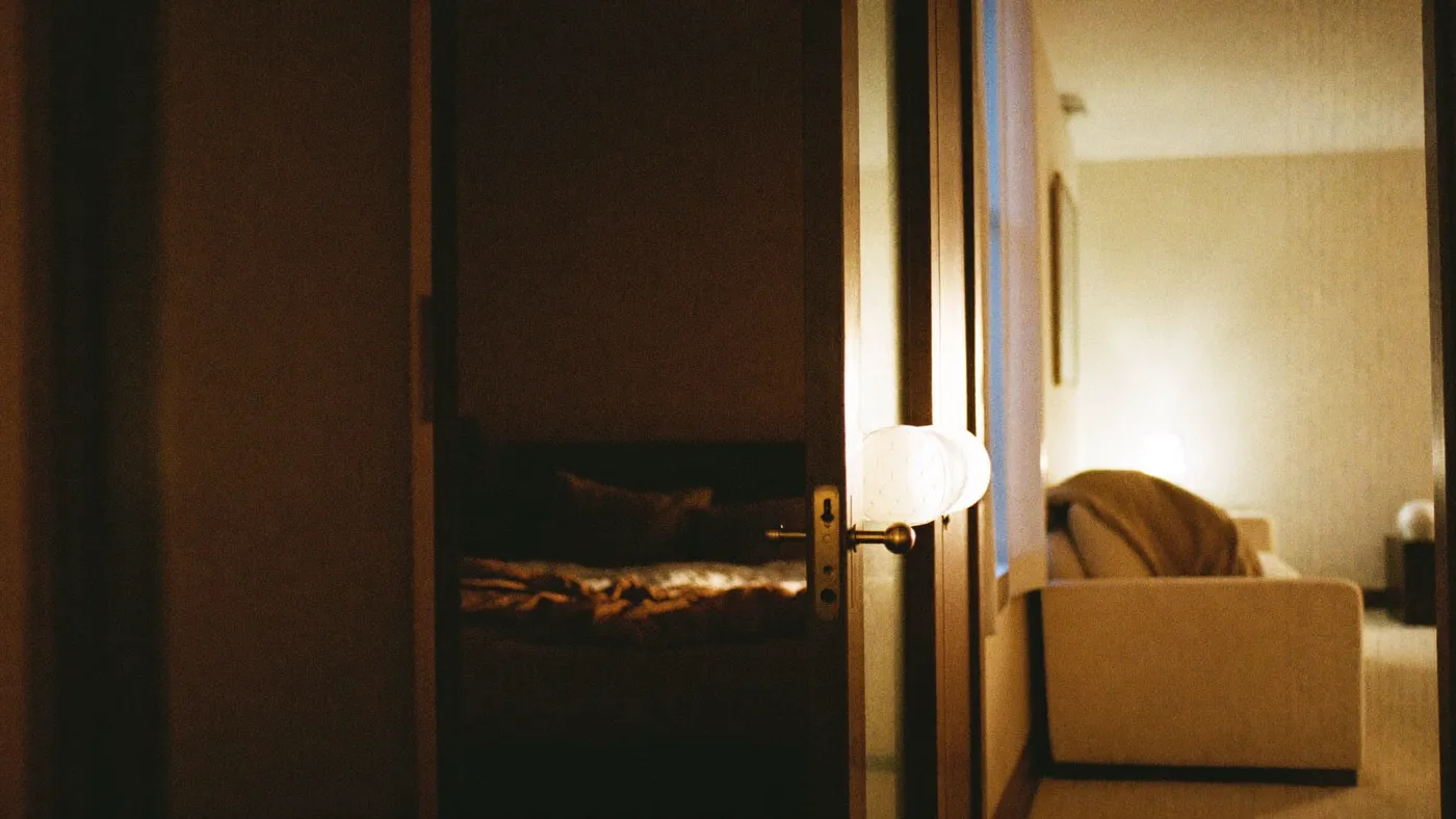 A dark bedroom doorway seen from a lit living room, a blanket folded on the sofa arm, warm lamp light