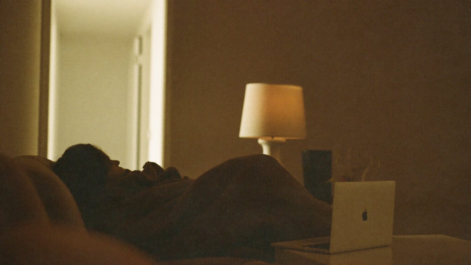 Person on a sofa late at night under a blanket, laptop closed on the coffee table, looking toward a dim hallway, soft lamp light