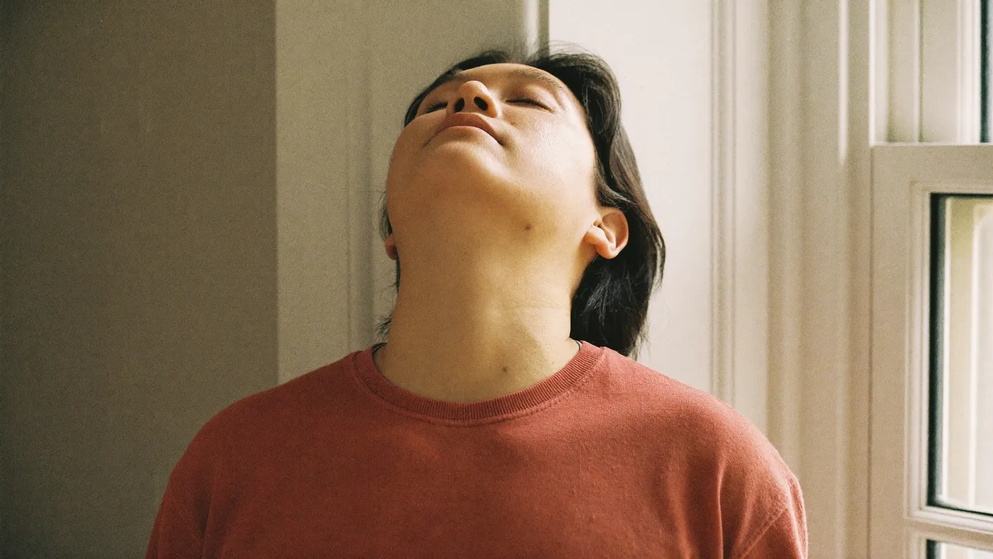Person standing at an open window with eyes closed mid-breath, soft city light behind them