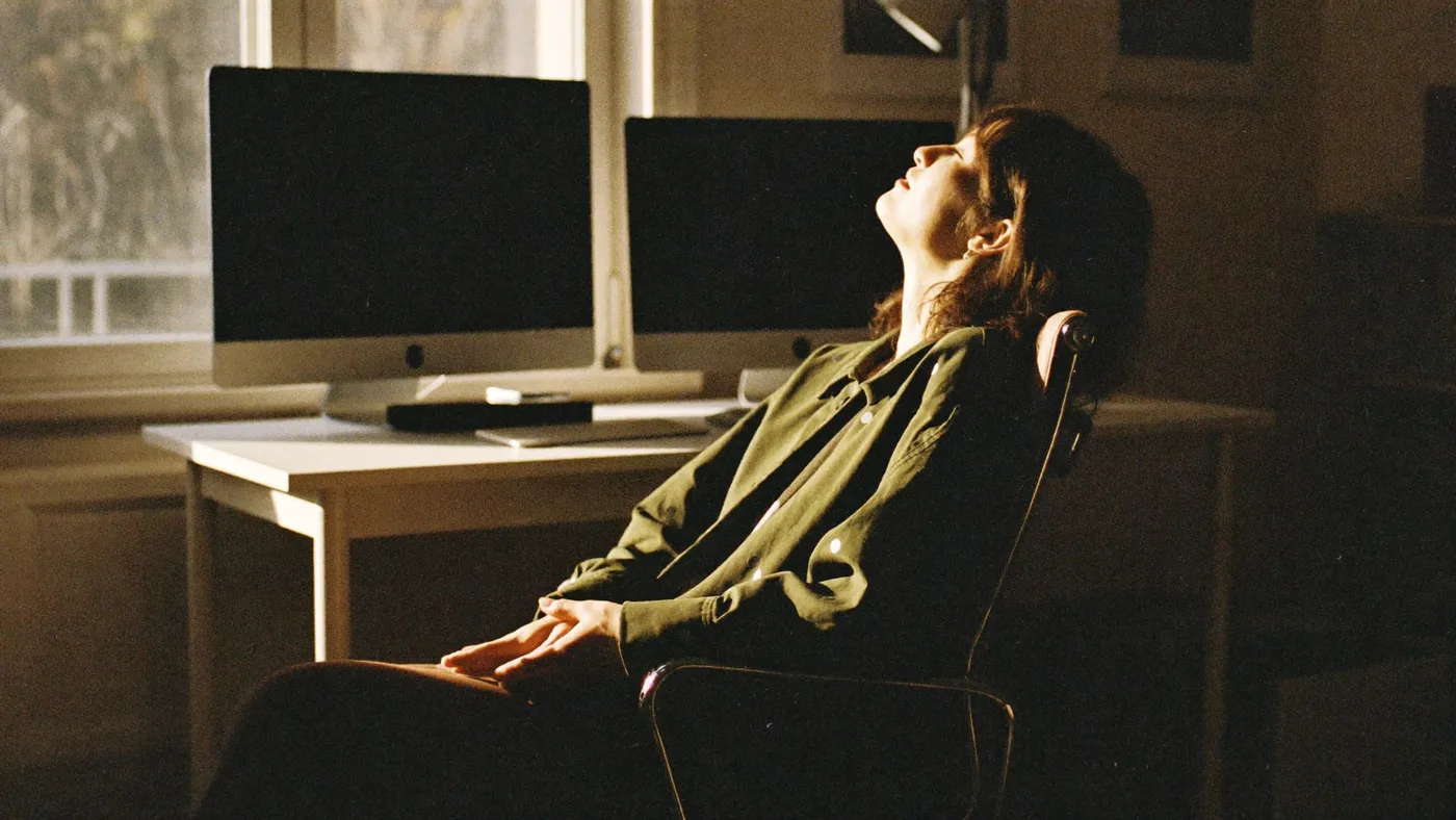 Person leaning back from a tidy desk with eyes closed and hands resting loose, warm office window light