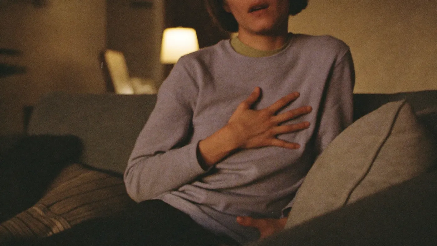 Person exhaling slowly with one hand resting on their chest, seated on a sofa at dusk, soft lamp glow