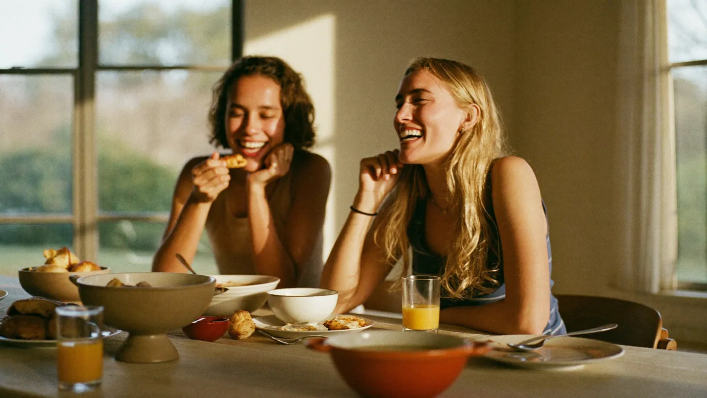 Two friends sharing breakfast at a sunlit kitchen table, candid easy laughter, warm homey tones