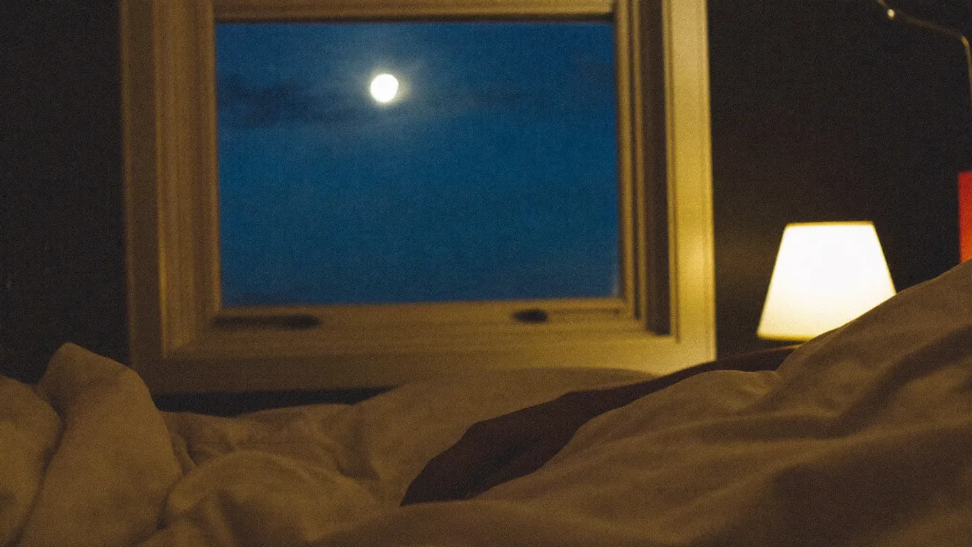 Bedroom at night, soft moonlight across rumpled sheets, a person's relaxed hand resting on the duvet