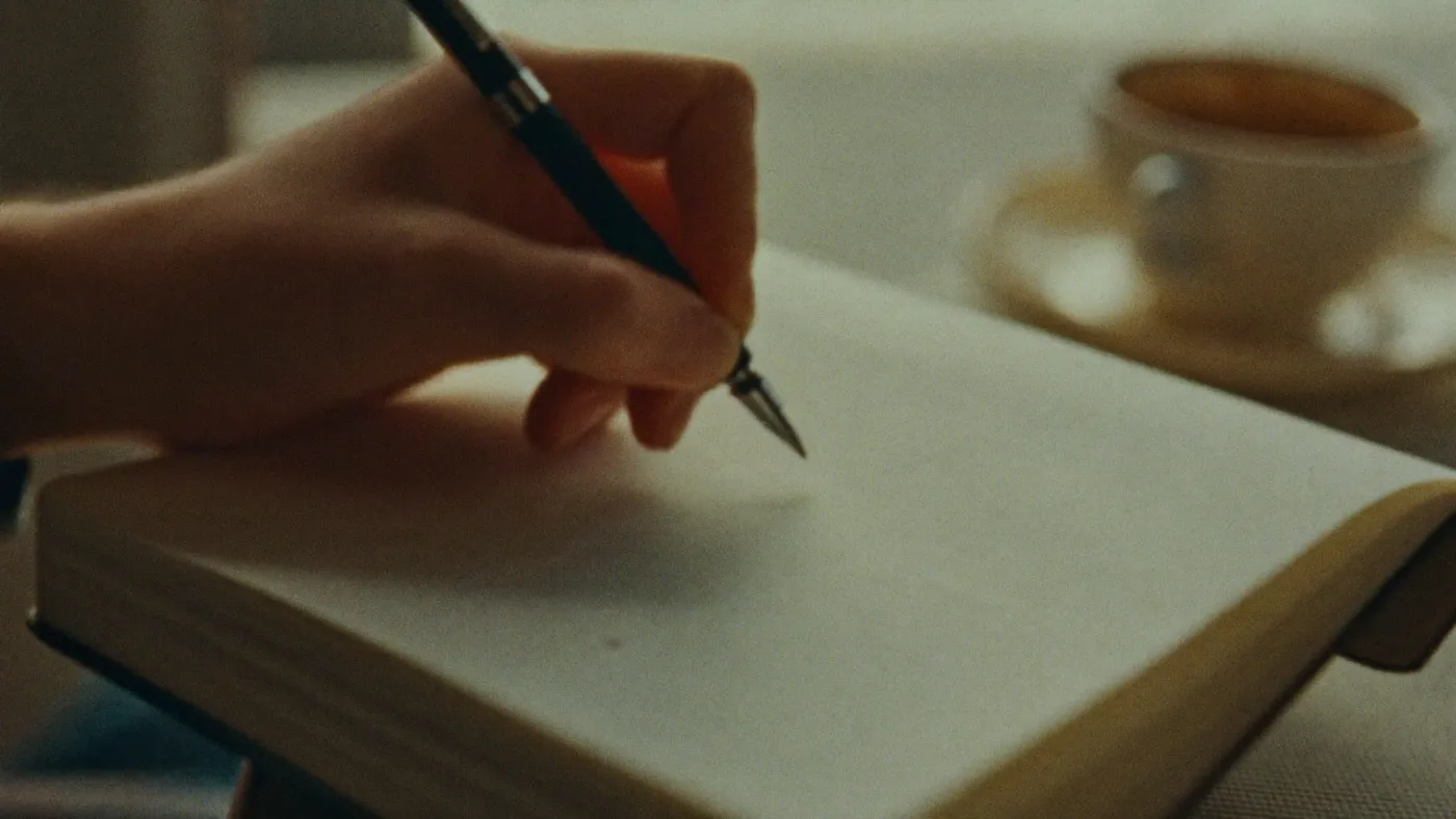 Close-up of hands writing in an open notebook with plain blank pages, cup of coffee nearby, morning window light