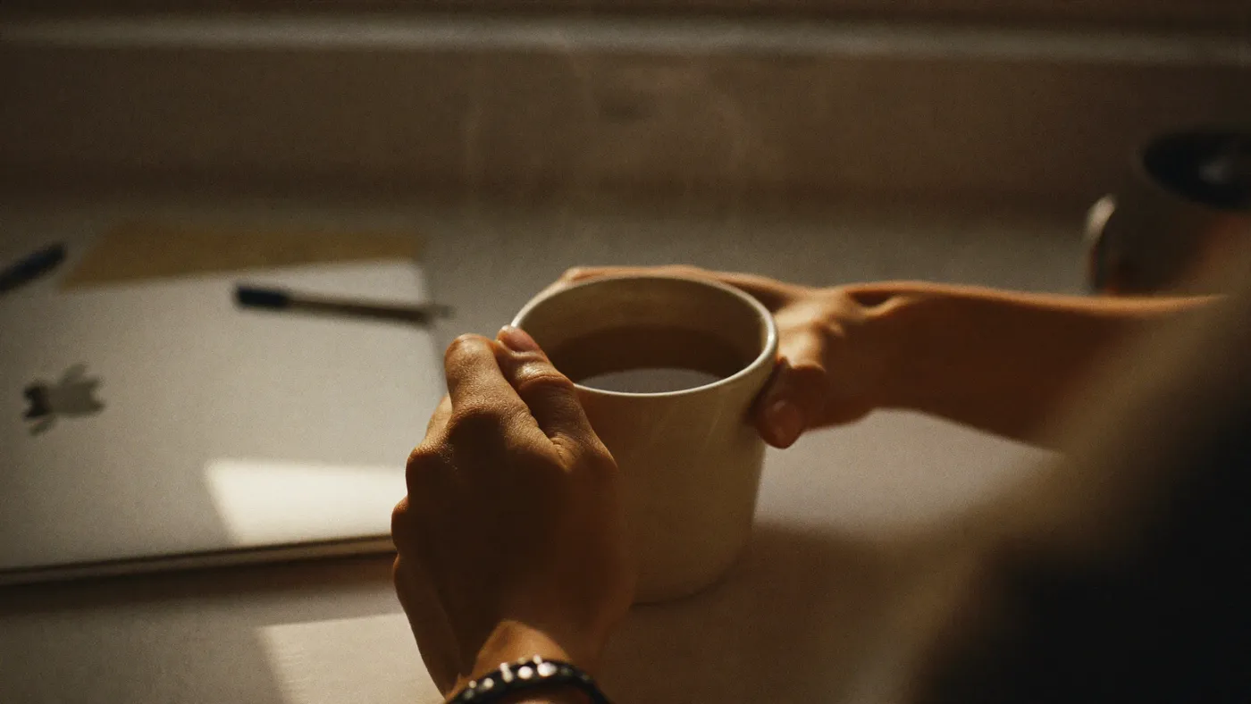 Close-up of hands around a warm mug on a desk beside a closed laptop, soft window light, quiet reset moment