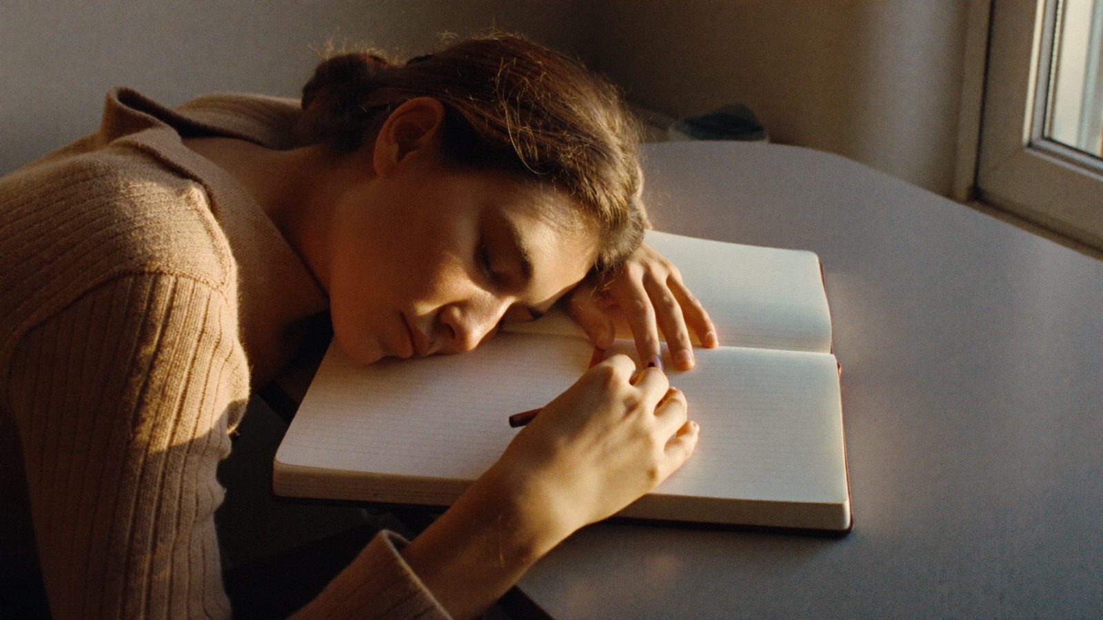 Person at a tidy desk by a window, pausing with eyes closed and a hand resting on an open notebook with blank pages, late afternoon light
