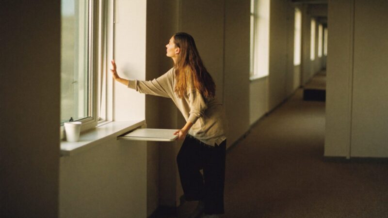Person pausing in a quiet office corridor by a tall window before a meeting, one hand on the glass, soft daylight