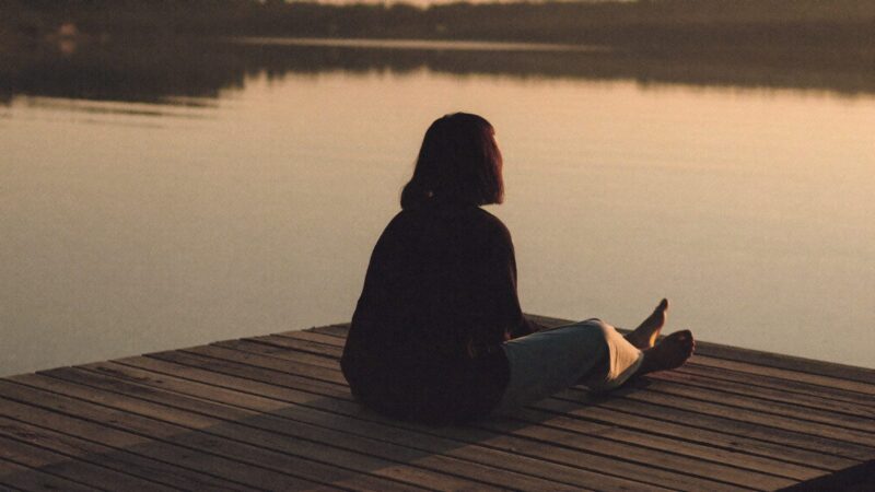 Person sitting alone on a wooden pier at golden hour with their feet over calm water, seen from behind