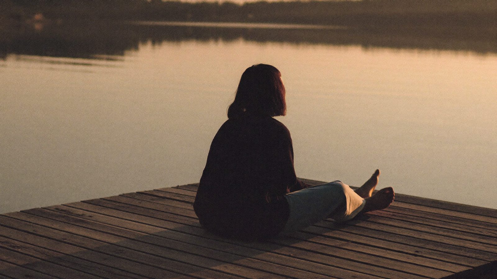 Person sitting alone on a wooden pier at golden hour with their feet over calm water, seen from behind