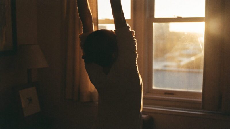 Person stretching their arms upward at a sunlit window first thing in the morning, warm light across the room