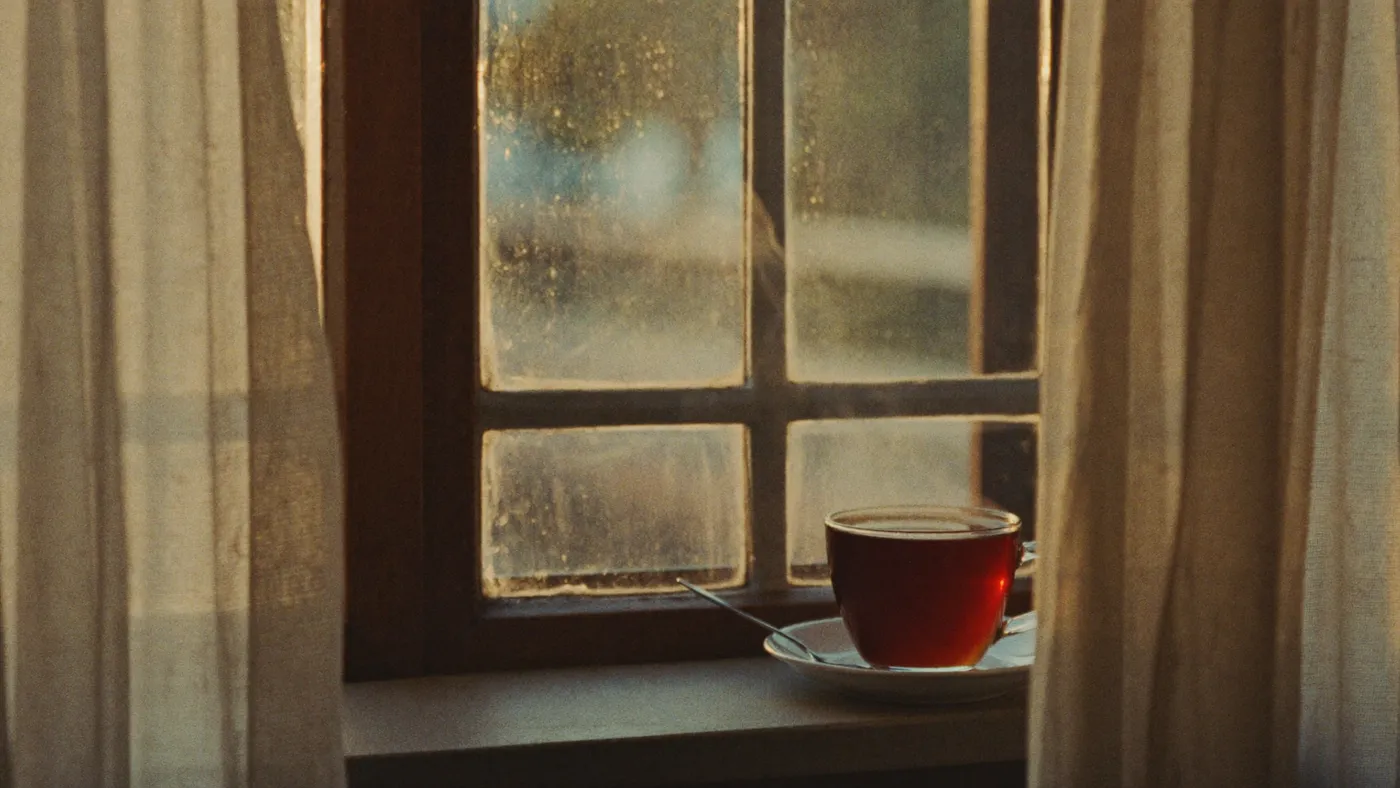 Steaming cup of tea on a windowsill at sunrise beside an open window with soft curtains moving
