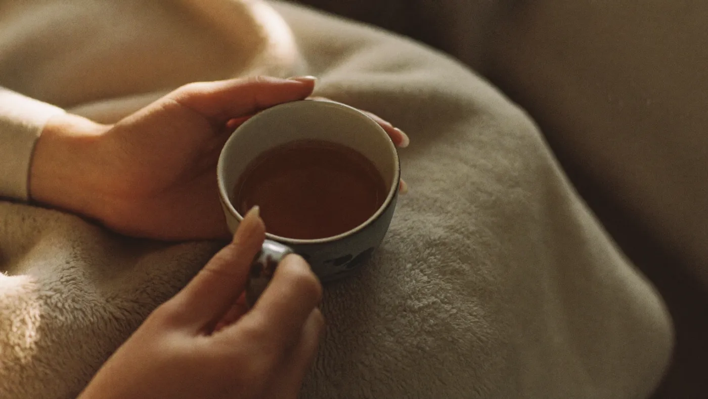 Close-up of hands cupping a small ceramic bowl of tea, soft blanket, warm quiet room, shallow depth of field