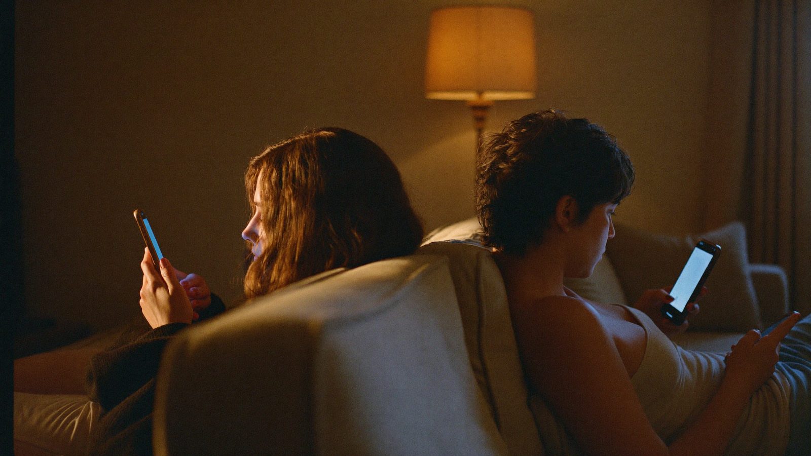 Two people on a sofa at opposite ends, both looking at their phones, warm lamp light, quiet distance between them