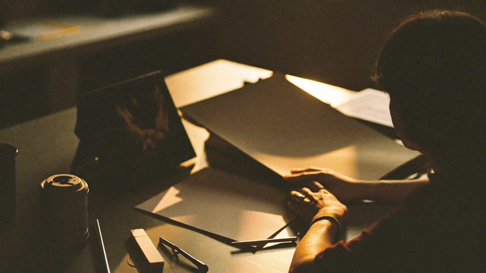 Person at a desk looking at a finished piece of work, hands resting, exhaling, warm late light, calm acceptance