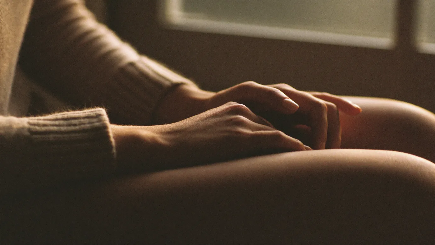 A hand resting gently over the other hand on a lap, soft knit sweater, warm window light, shallow focus