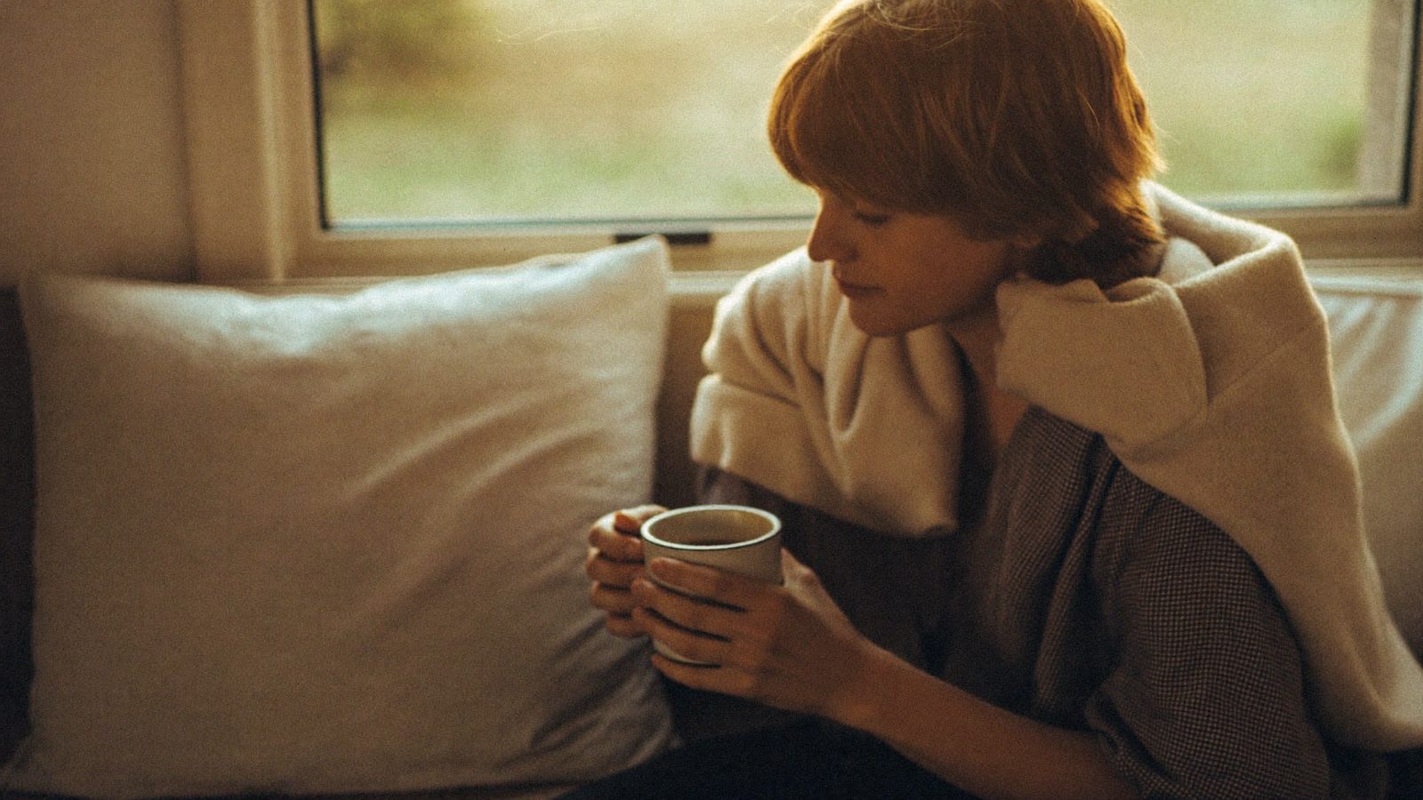 Person sitting on a window seat with a blanket around their shoulders, holding a mug with both hands, kind soft expression, morning light