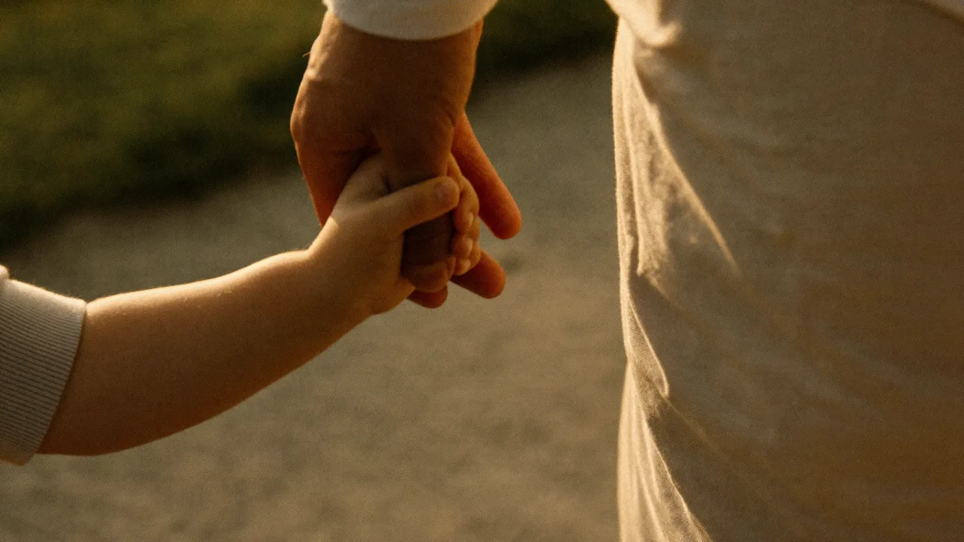 A child's hand holding an adult's finger while walking on a sunlit path, close-up, warm light