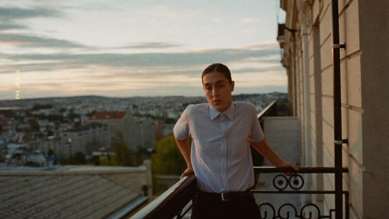 Person standing on a balcony at dusk with hands on the railing, breathing out, city softly blurred below