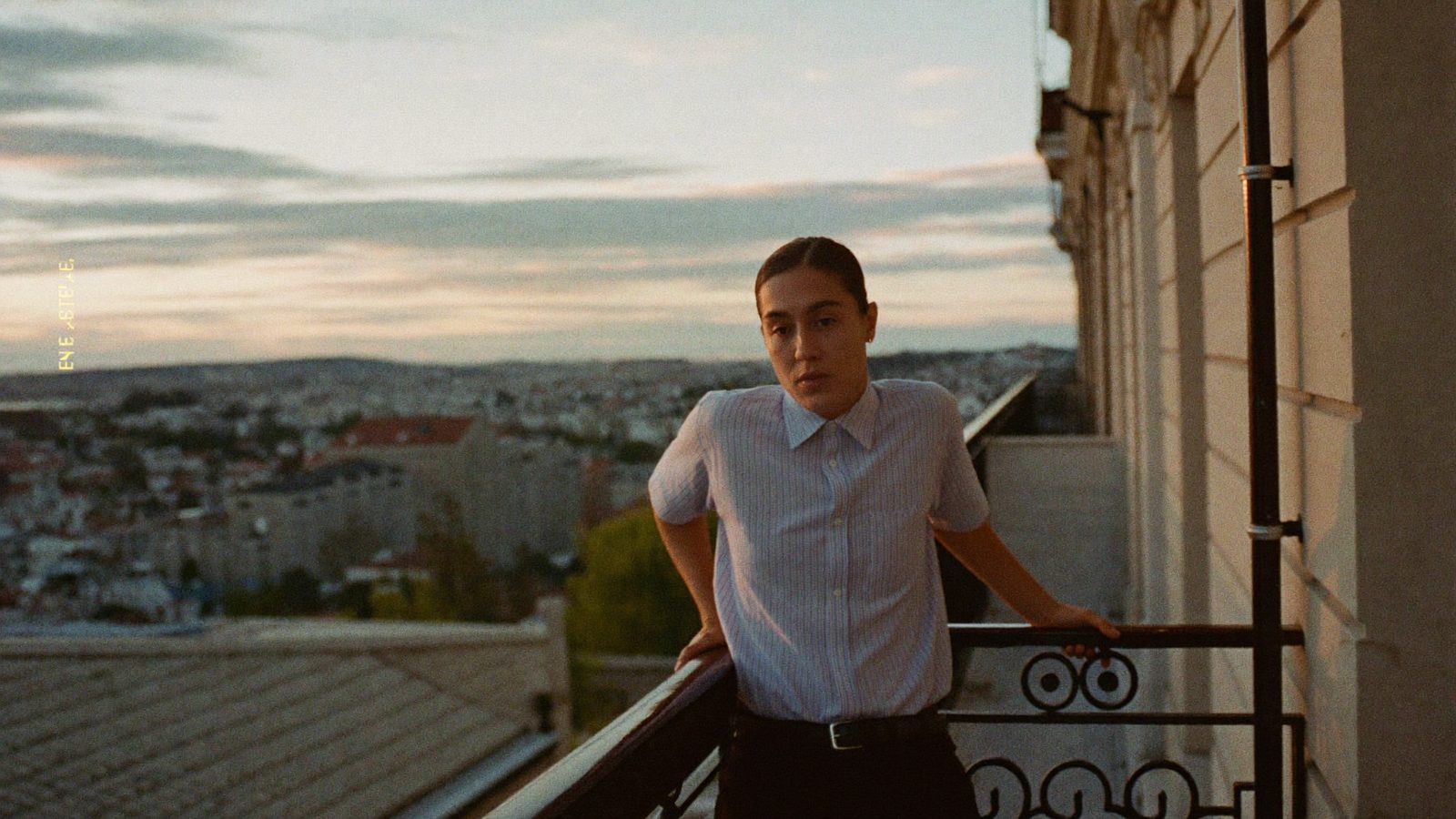 Person standing on a balcony at dusk with hands on the railing, breathing out, city softly blurred below