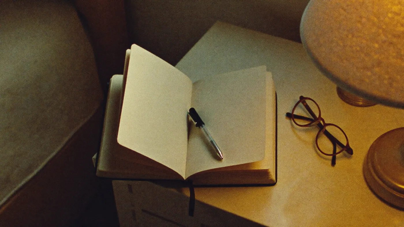 A small notebook with blank pages, a pen and reading glasses on a bedside table, warm lamp light