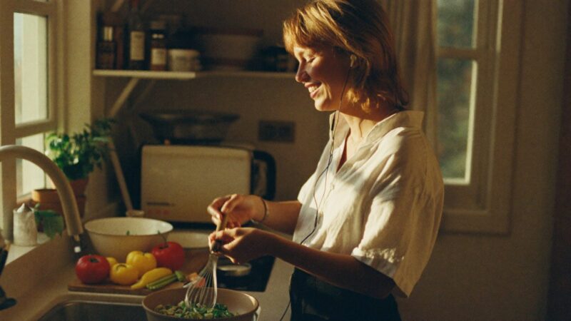 Person listening with earbuds in a sunlit kitchen while cooking, smiling at something they hear, candid
