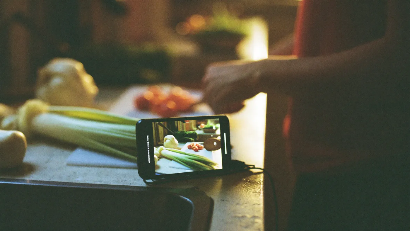 A phone propped on a kitchen counter with a soft blurred glow while a person chops vegetables, warm kitchen light